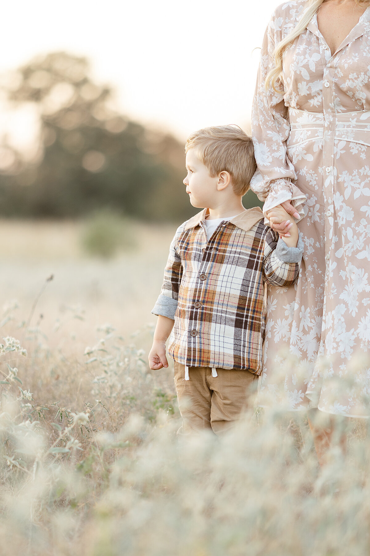 A little boy standing in a grassy field while he holds his moms hand.