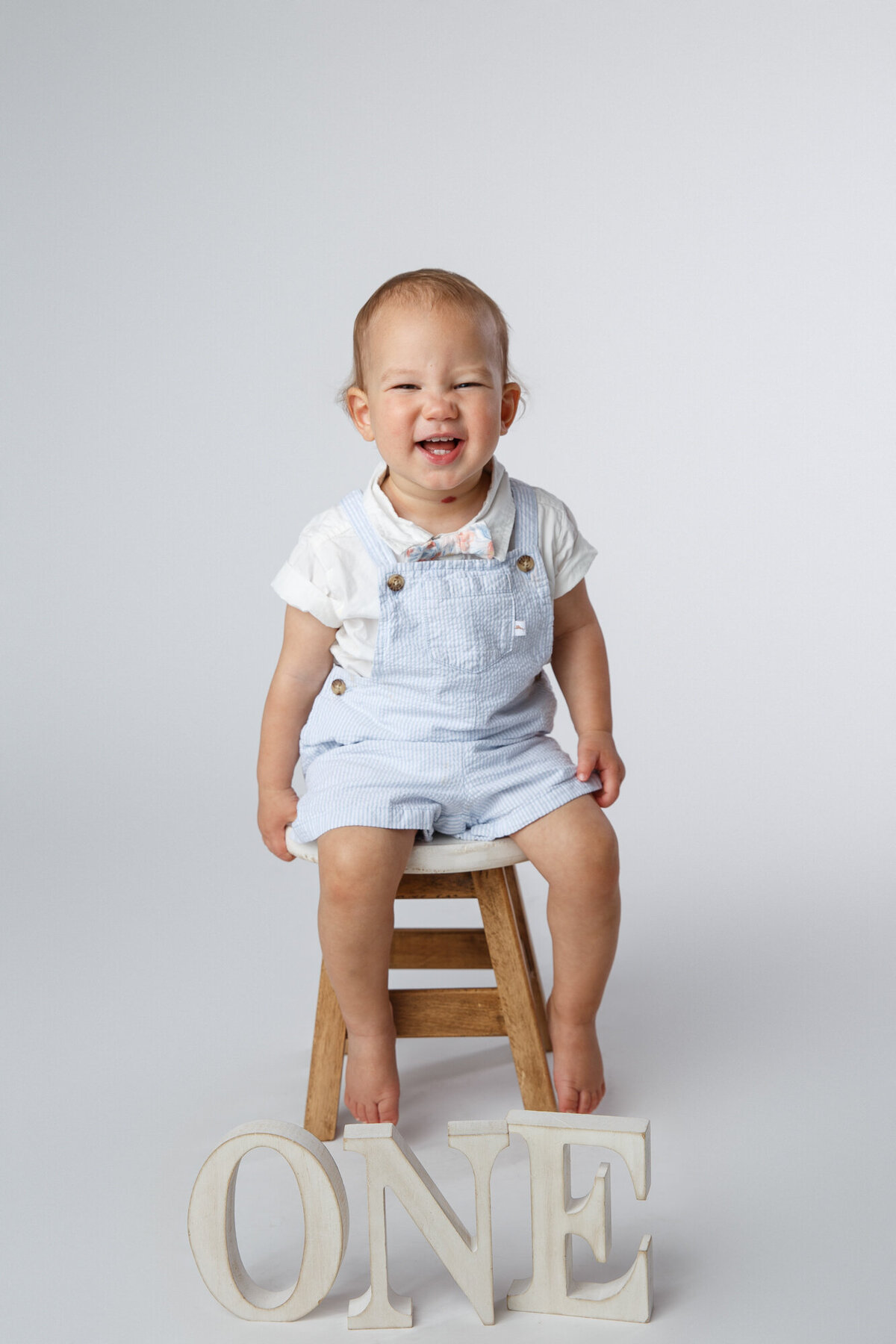 Cute one year old baby sitting on a stool smiling with the letters spelling one in the foreground