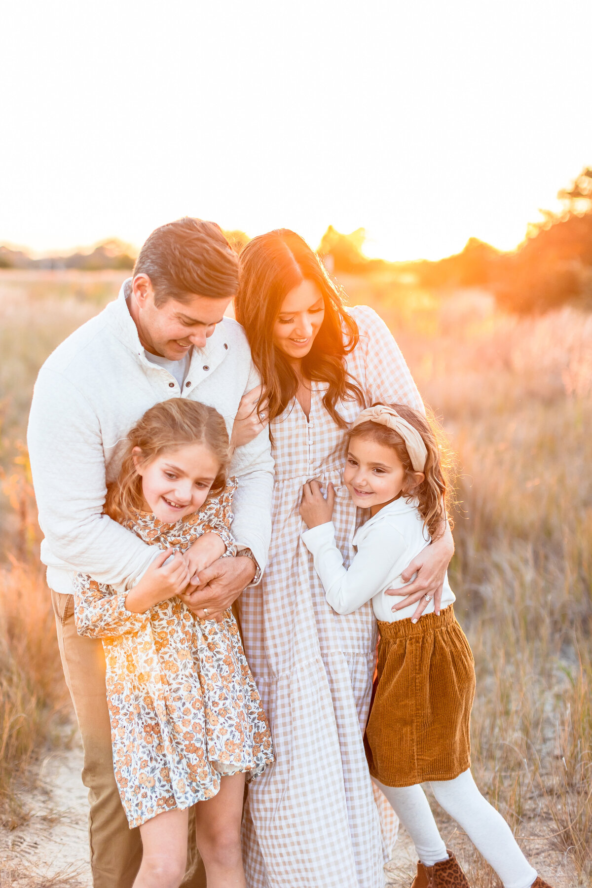 A loving family of four embraces in a field of golden light at sunset in Virginia Beach, their laughter and warmth glowing in the golden hour as they soak in the moment together.  