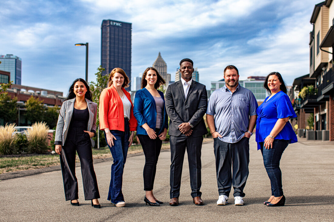 A group of six professionally dressed people standing in a line outdoors with a city skyline visible in the background. The Pittsburgh skyline can be seen with tall buildings and what appears to be the state capitol dome. The group is wearing business attire in various colors.