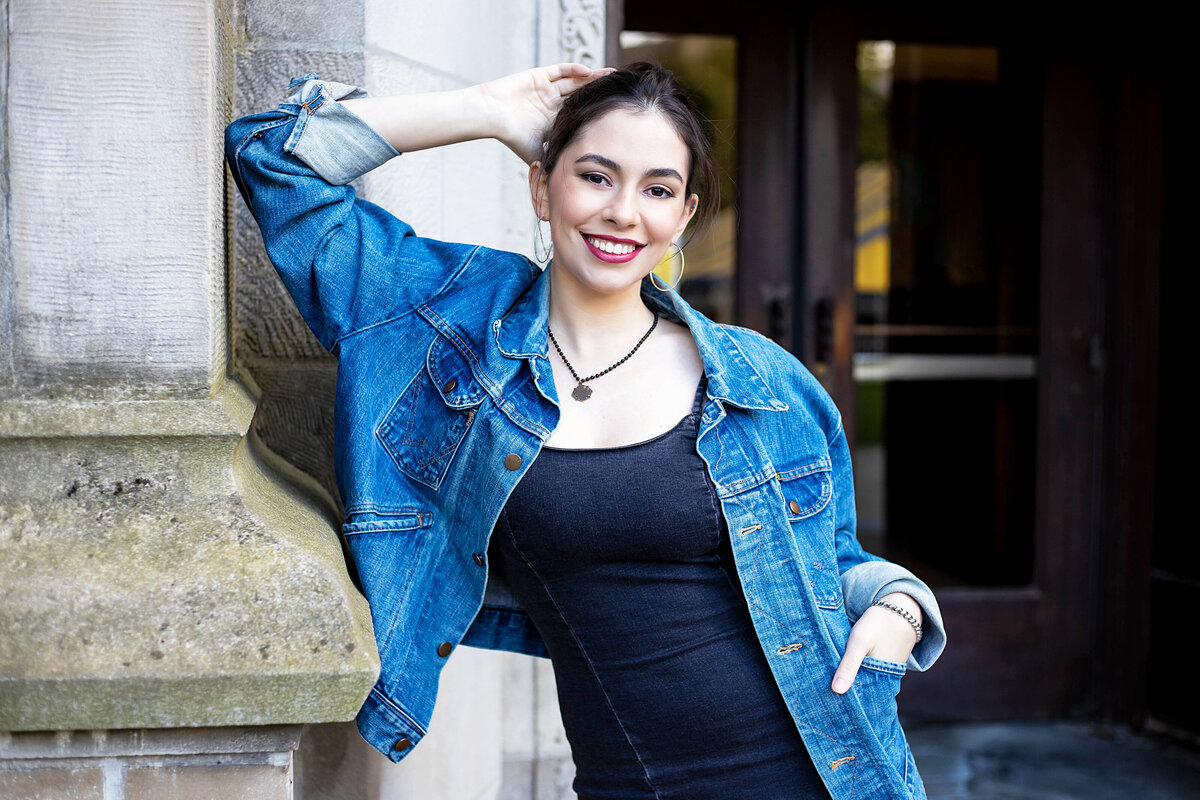 Ann Arbor U of M Law Quad Senior Photoshoot | Ann Arbor Michigan Senior Photographer, Senior girl with black hair in blue jean jacket and black jean dress leaning on brick in front of the entrance to the library