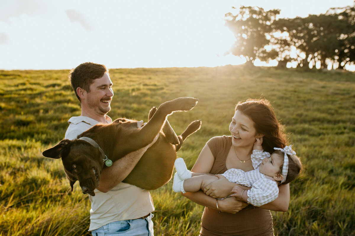 Parents share a humorous moment, mom carrying the baby and dad holding the Labrador similarly.