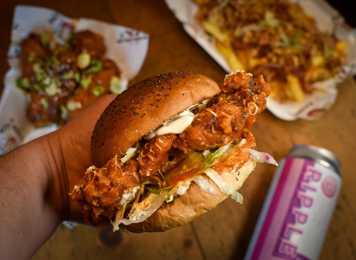 Hand with wrist holding a chicken burger with loaded fries and beers in the background