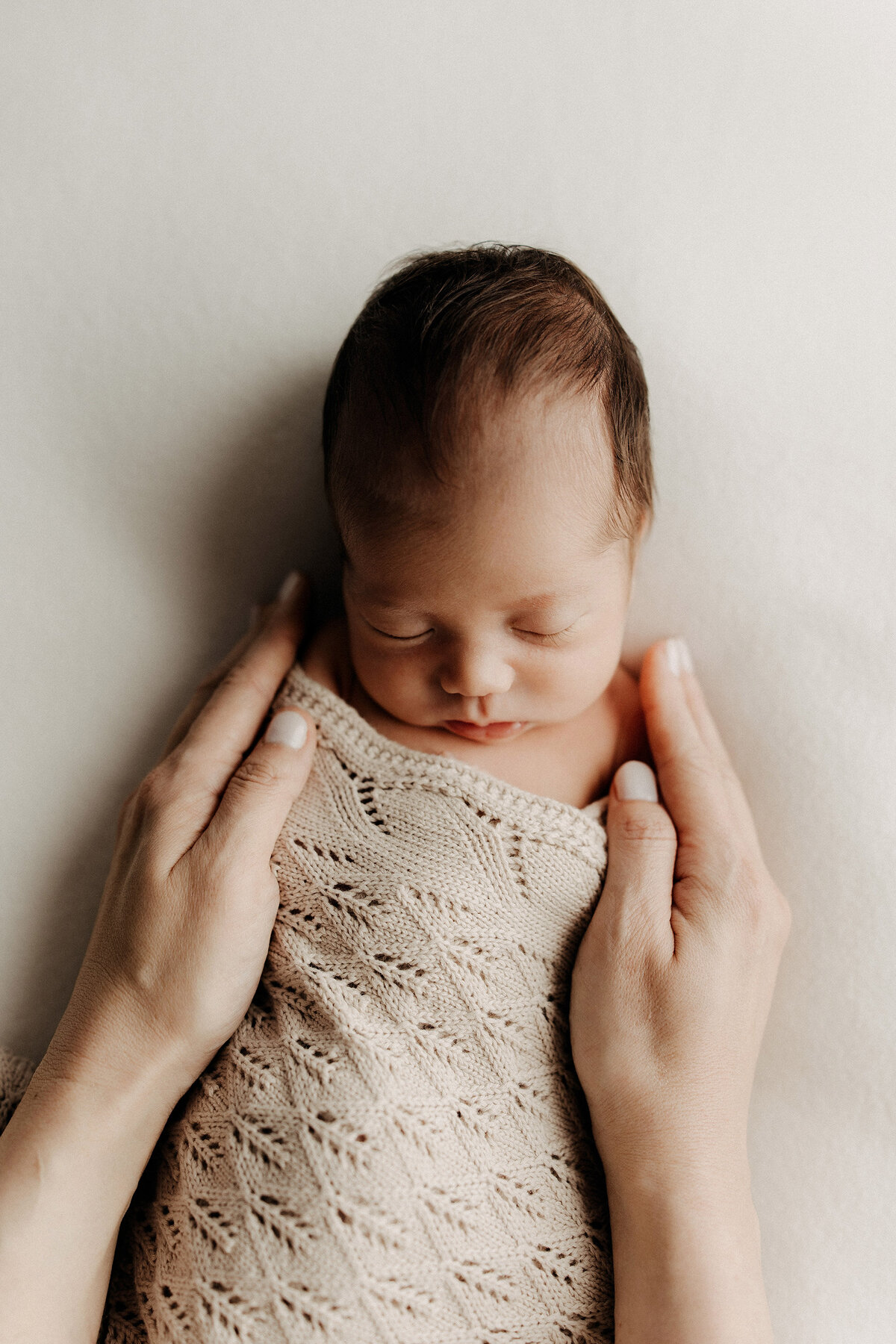 Newborn baby sleeping on a cream surface, wrapped in a beige knit blanket, with two gentle adult hands cradling them from above.