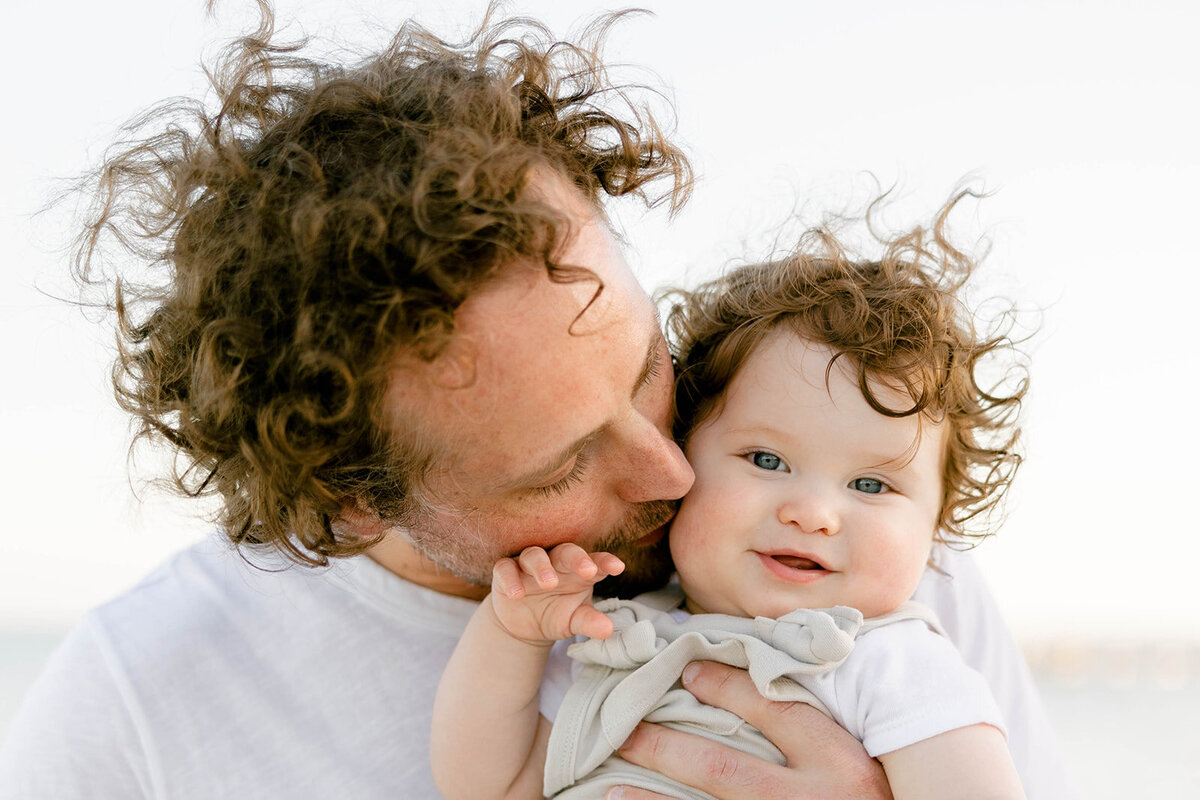 A father kisses his curly-haired baby, sharing a warm moment during a Florida Keys family session. Captured by Claudia Amalia Family Photography.