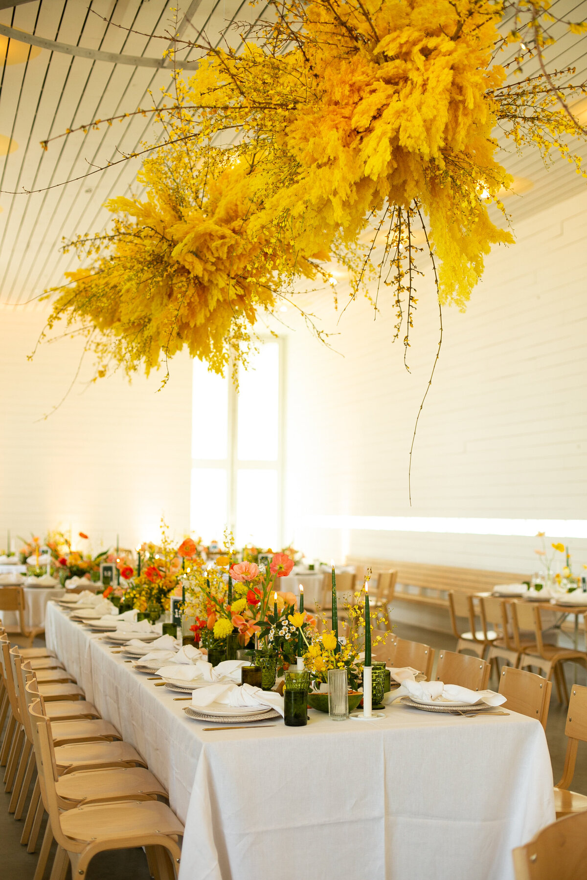 Floral ceiling installations in bright yellow hung above tables with reds and orange florals and green glasses.
