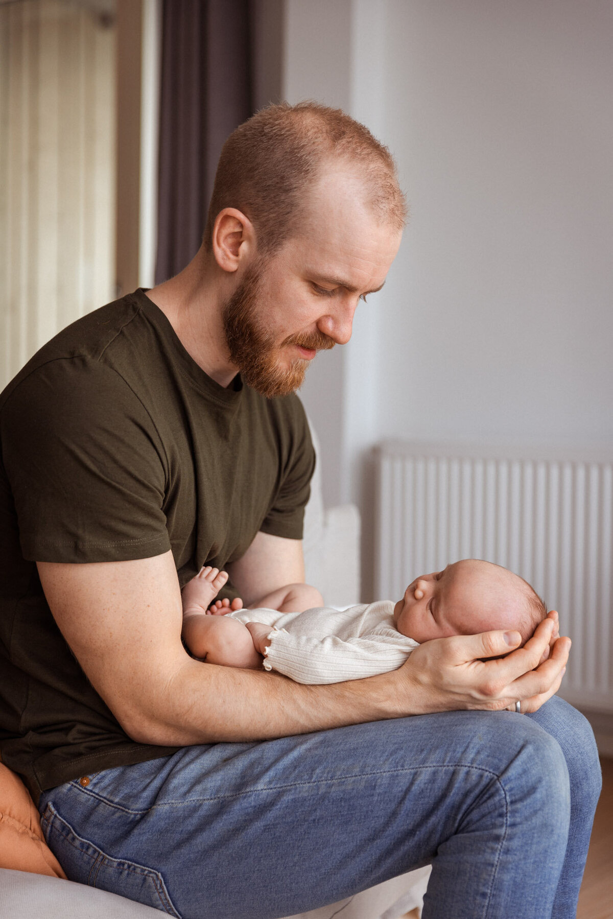 Livsstilsfoto av far i profil, far holder babyen sin på knærne sine, babyen ligger i armene hans med hodet i hendene hans.