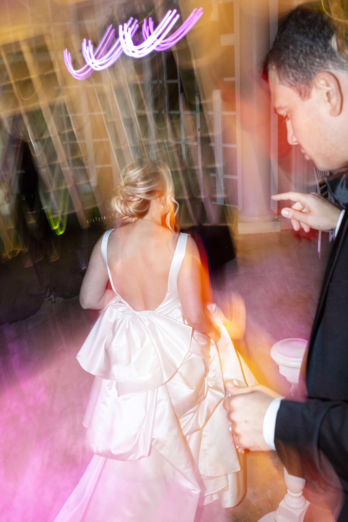 A woman in a white dress with a large bow at the back is dancing. She is surrounded by colorful lights and blurry motion. A man in a suit is dancing beside her. The scene captures a joyful moment at an event or celebration.