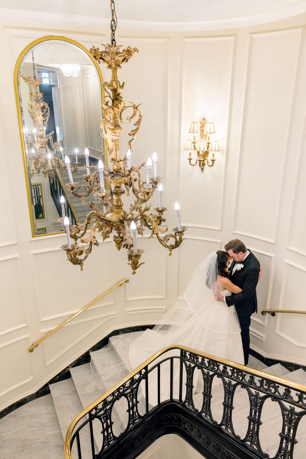 A bride and groom embrace on a grand staircase. The bride wears a flowing white gown and veil, and the groom is in a black suit. A large ornate chandelier hangs above, and a mirror reflects part of the elegant staircase.
