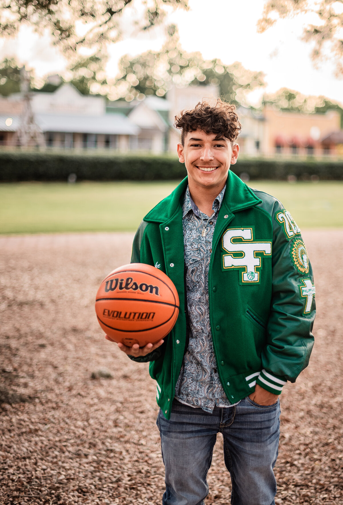 A senior boy wears his letterman and holds a basketball while he smiles for a picture.