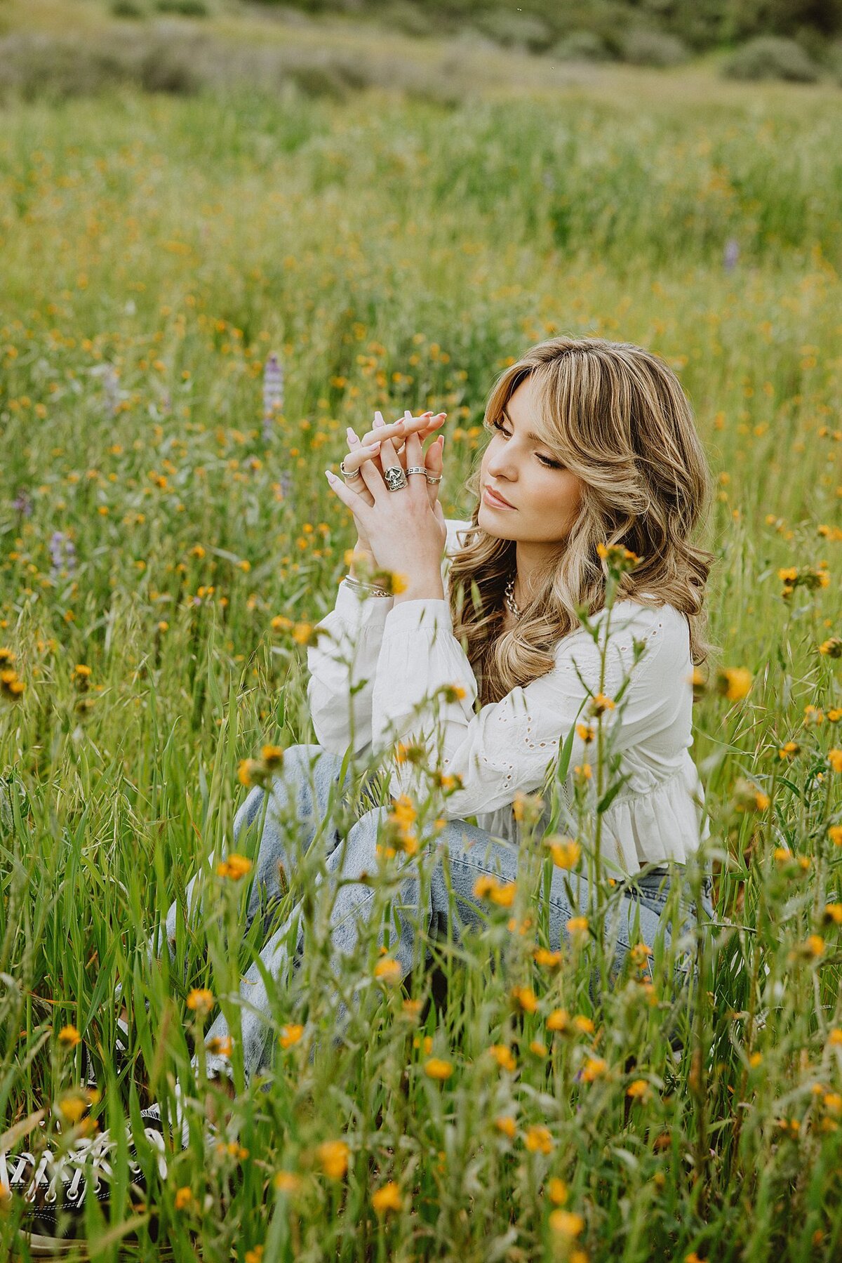Teen girl in a white blouse and jeans sitting in a blooming wildflower meadow, gazing dreamily at her hands.