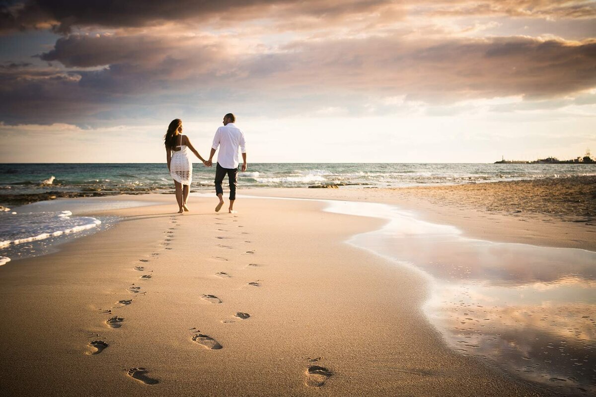 Couple walk hand in hand on the beach at sunset leaving footprints as they walk