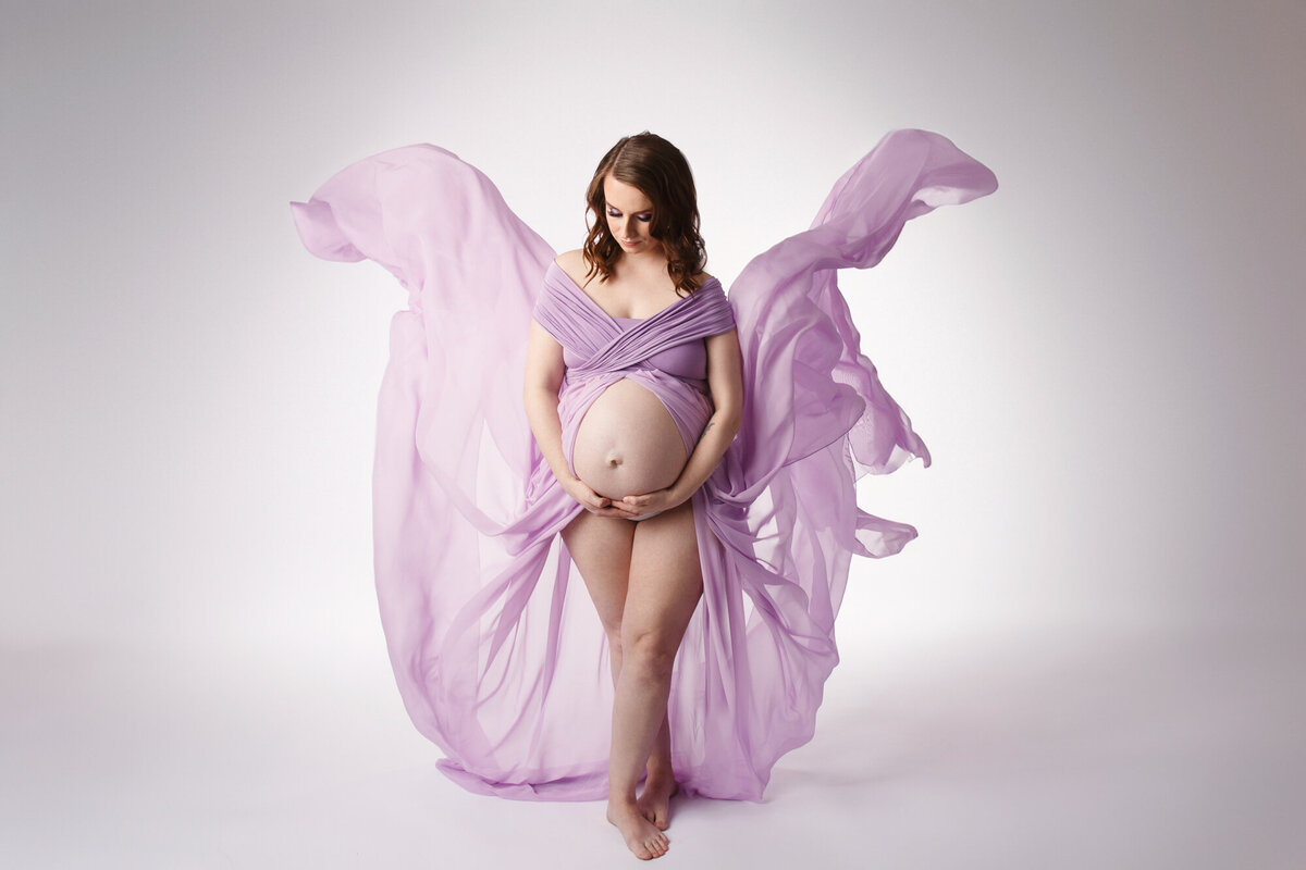 Beautiful motherhood portrait of woman wearing a purple gown looking down at her baby bump while the material of her gown flows upwards behind her