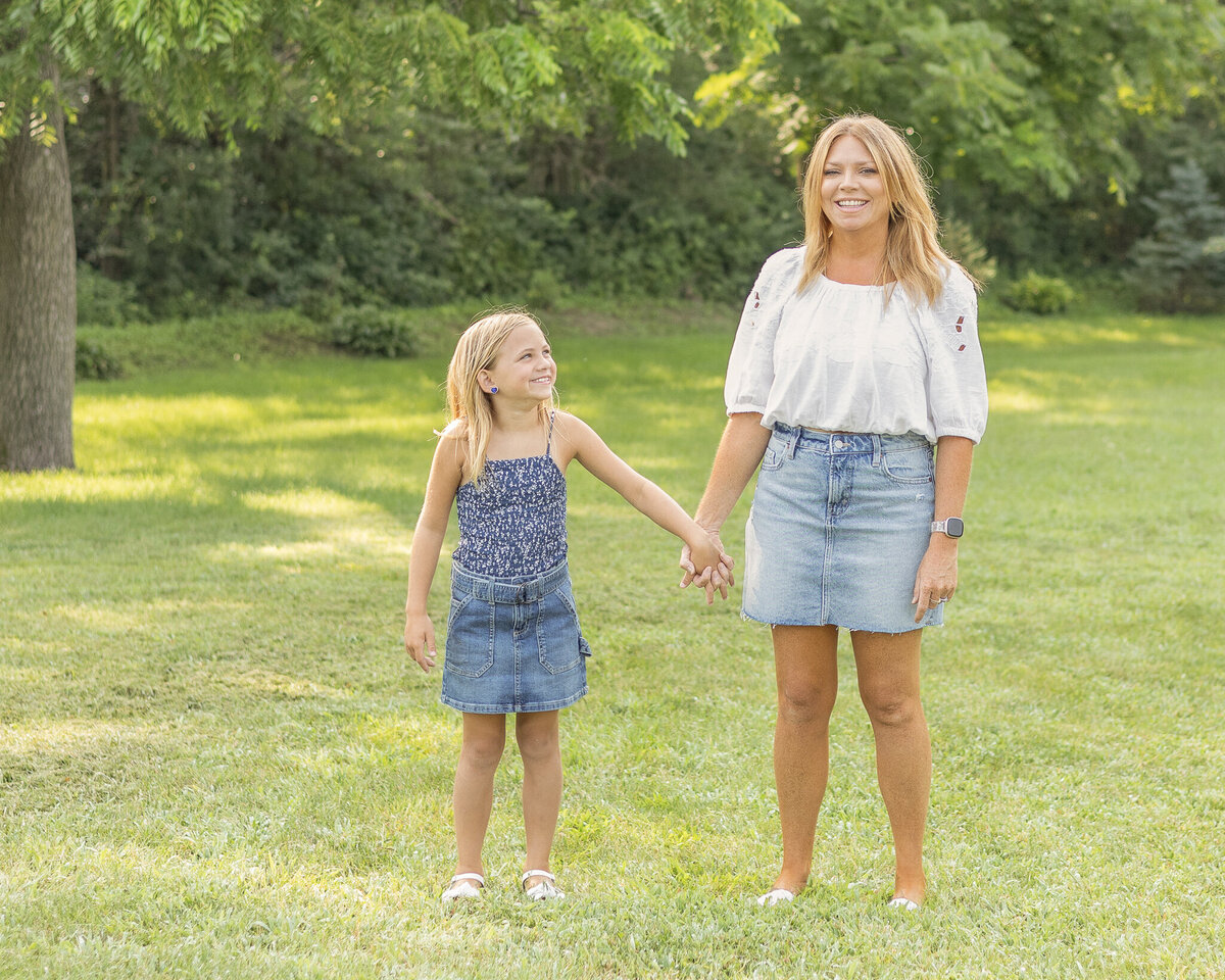 Mom and youngest daughter standing in the grass with a tree line behind them, holding hands.  Youngest daughter is looking at mom smiling, wearing a blue jean skirt, and dark blue and white tank stye shirt.  Mom is wearing a jean skirt, white half sleeve shirt, smiling at the camera.