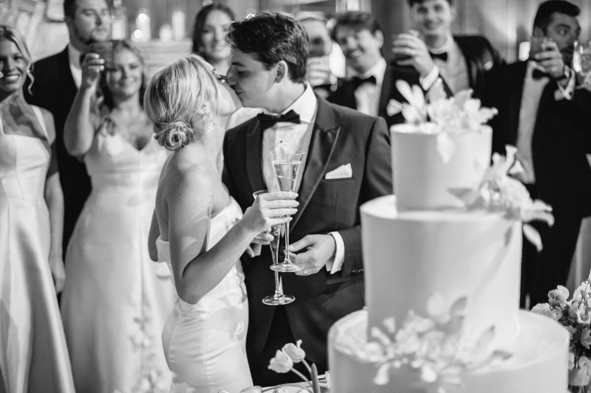 Black and white photo of the bride and groom toasting and kissing during the cake cutting at their Highlands, North Carolina wedding at Old Edwards Inn.
