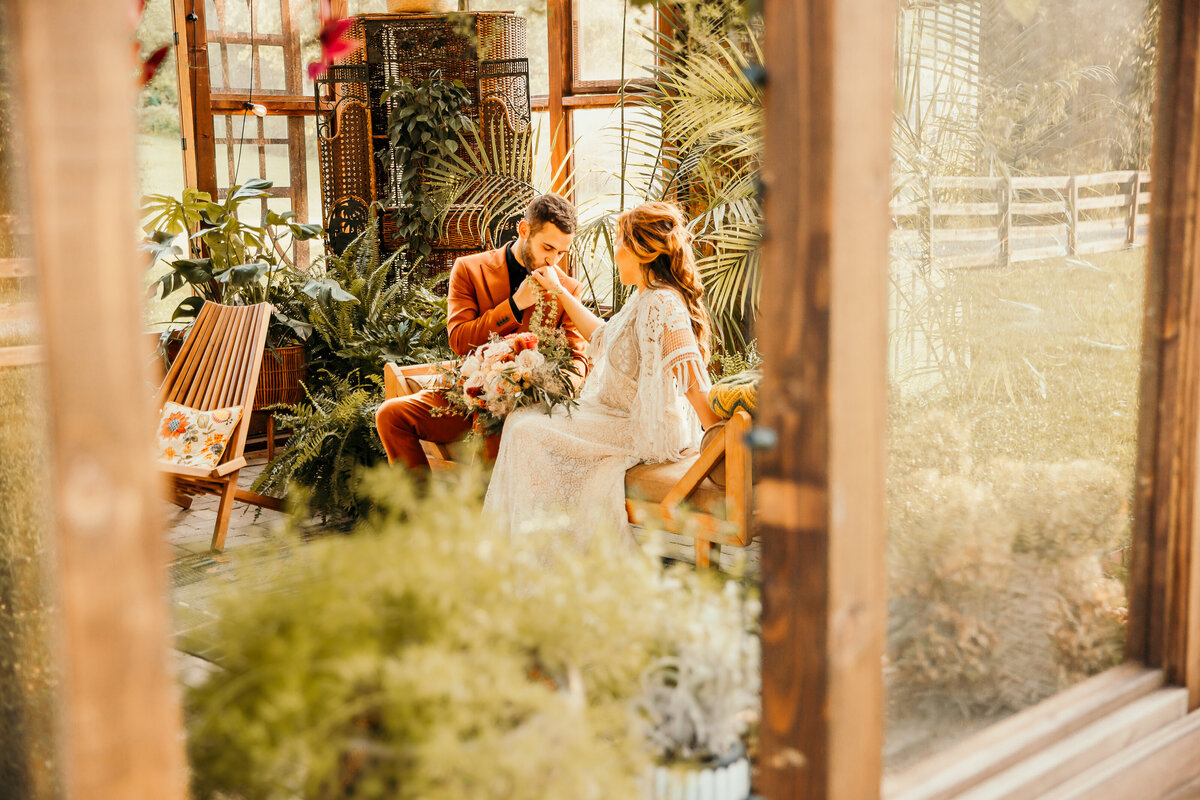 groom kissing bride's hand in greenhouse seen through open window