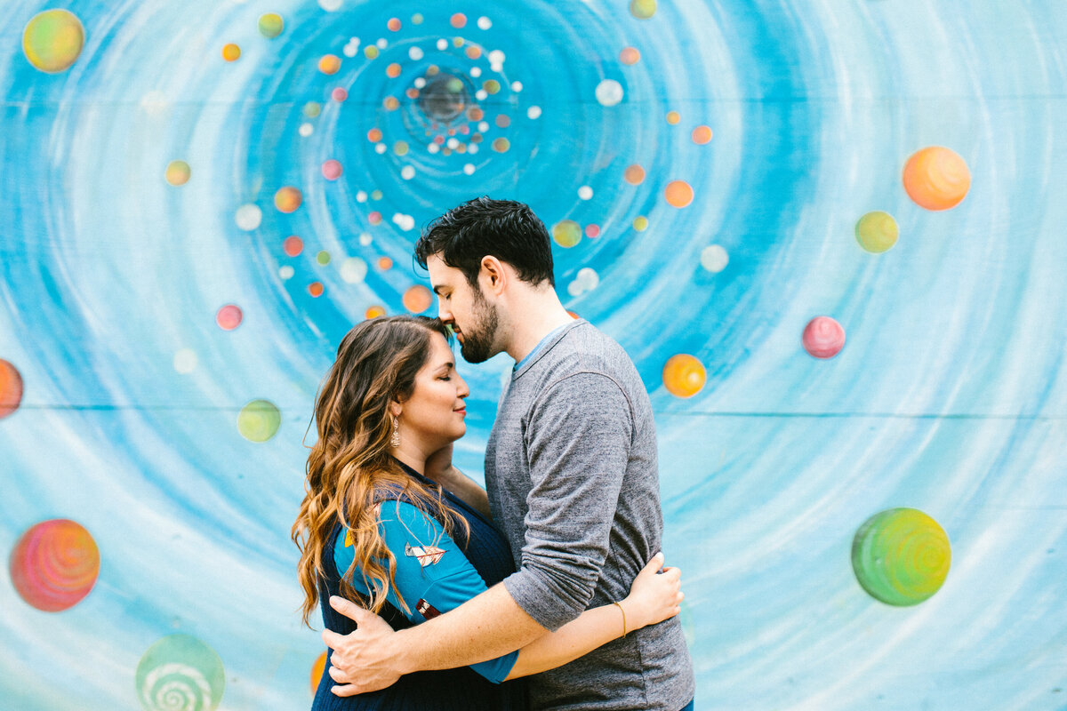 A couple holding each other close in front of a swirling, colorful mural during their engagement session in the Bishop Arts District in Dallas, Texas. The woman on the left is wearing a blue top with a dark blue vest while the man on the right is wearing a grey shirt.