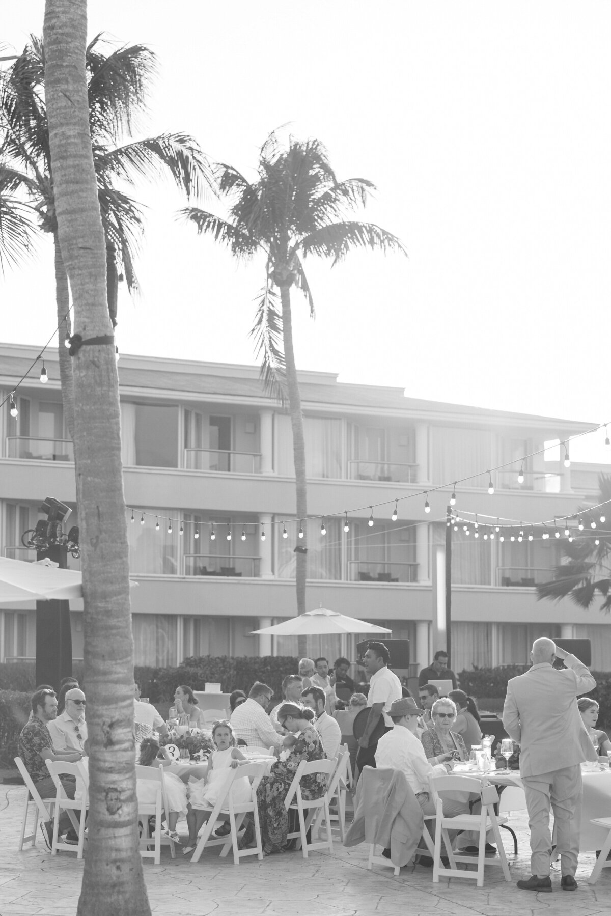 Guests dine outdoors at a Cancun resort wedding reception, with palm trees and bistro lights overhead.