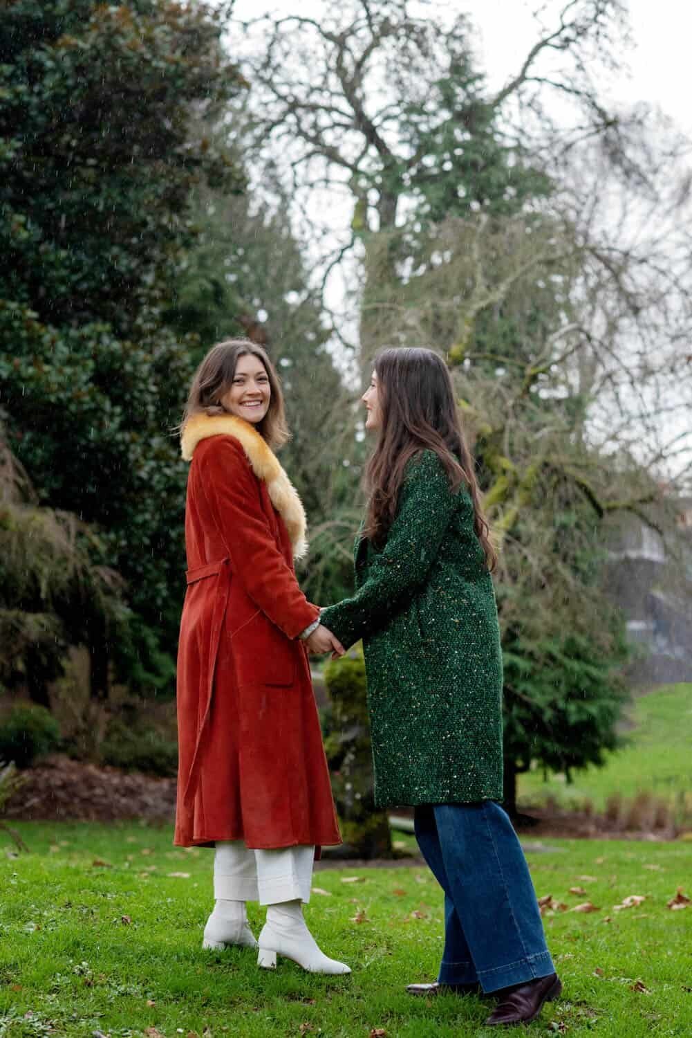 a woman in a vintage orange coat holds hands with a woman in a dark green coat