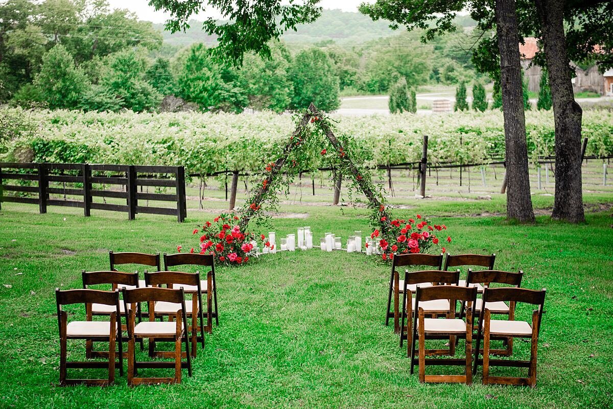 Brown wood garden chairs with ivory seat cushions are set up for a wedding ceremony at Arrington Vineyard with a triangular dark wood arbor covered in green vines and large centerpieces of red roses at the base. A long row of white pillar candles are set up at the bottom of the triangular wedding ceremony arbor facing the trees and vines of the vineyard at Arrington.