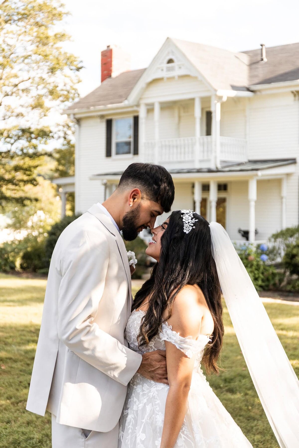 A newlywed couple stands in front of a historic white Victorian-style home in Nashville, forehead to forehead with soft smiles. The groom, dressed in a light cream suit, gently cradles the bride’s waist as she looks up at him with adoration, her long dark hair cascading over her shoulders