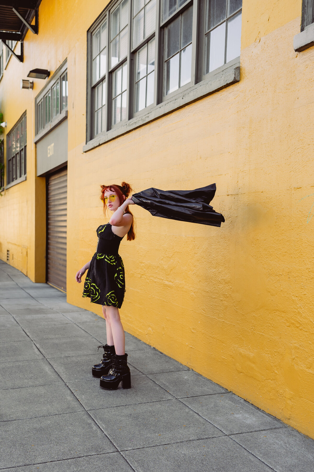 Woman flipping her jacket against a bright yellow building – street-style brand photo by Mewphotovideo