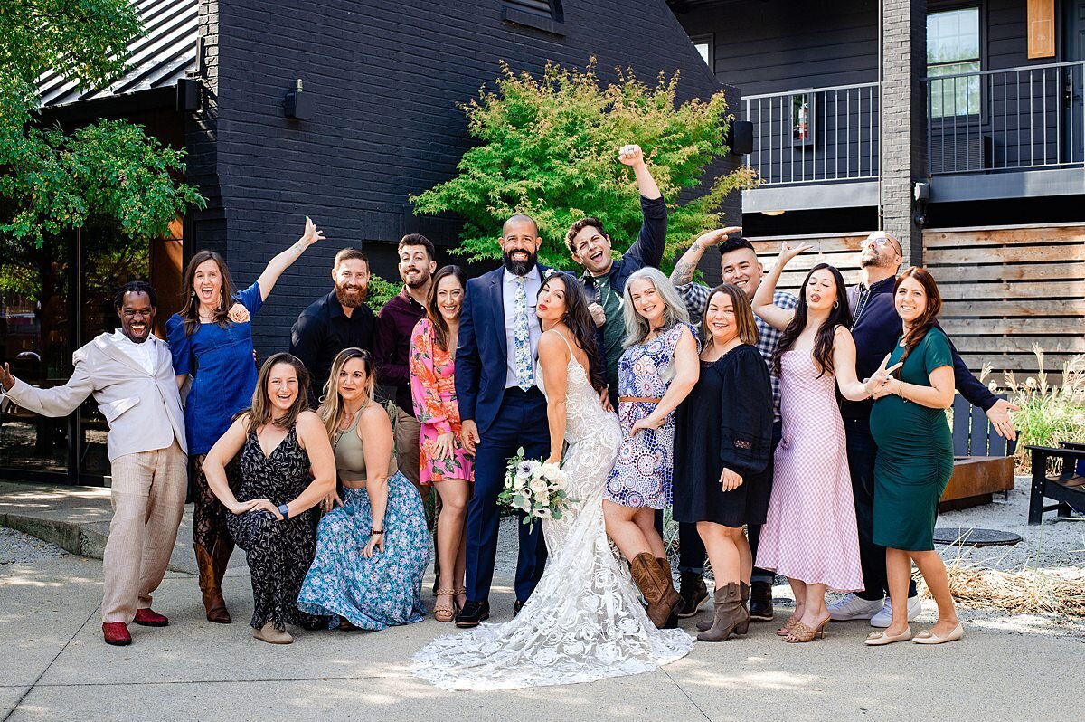 The bride, wearing a long lace wedding dress, holding a white bridal bouquet leans into the groom who is wearing a dark grey suit with a blue floral tie. On either side of them their family and friends strike silly poses as they celebrate their Nashville micro wedding at BODE Nashville.