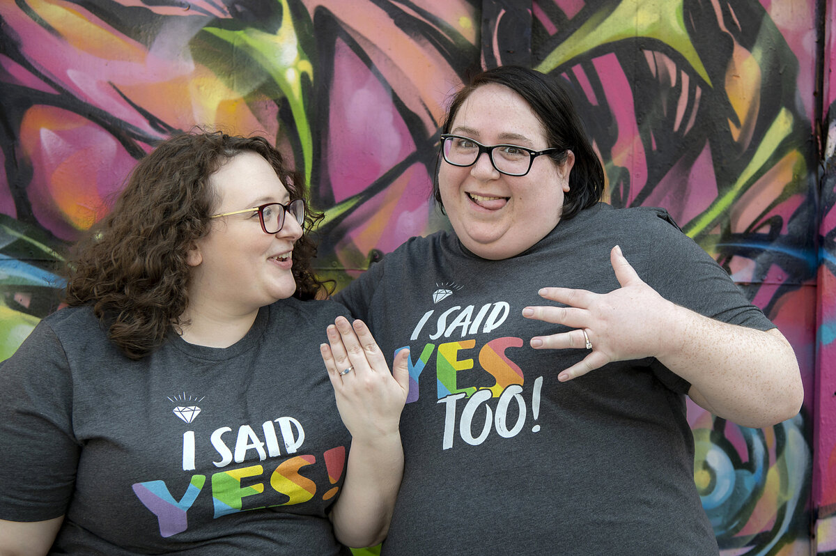 A couple smiling in matching tshirts showing off engagement rings 