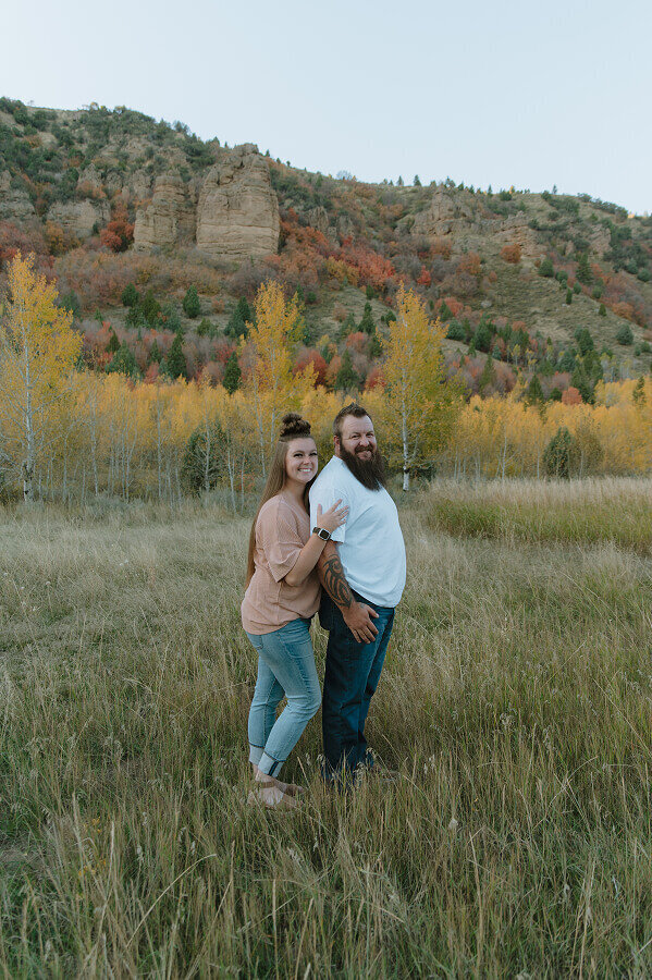 A wife holds her husband's shoulder from behind as they both smile for the camera. They're outside with lots of beautiful fall-colored foliage behind them.