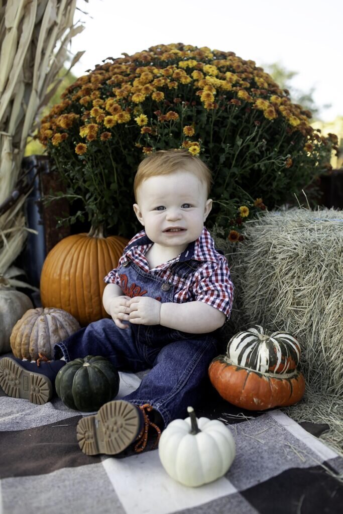 One-year-old child sitting on a bale of hay in the pumpkin patch, surrounded by pumpkins, with a big smile on their face