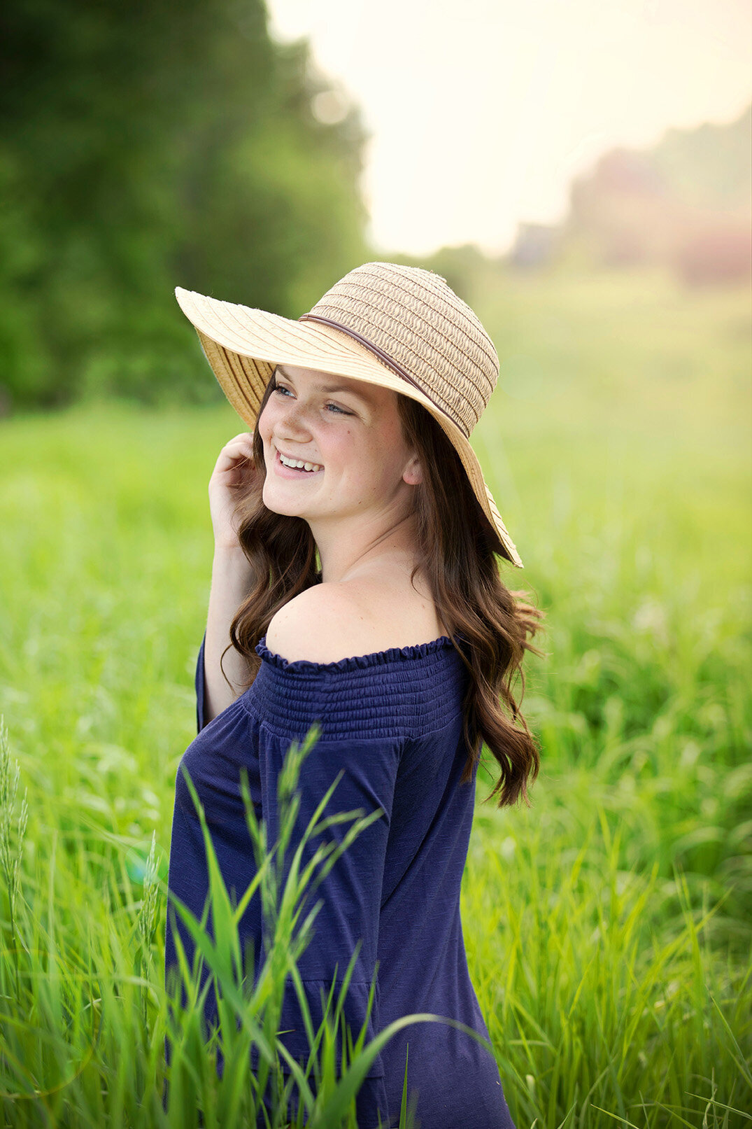 Senior girl wearing blue shirt and floppy hat standing in field of tall grass smiling.