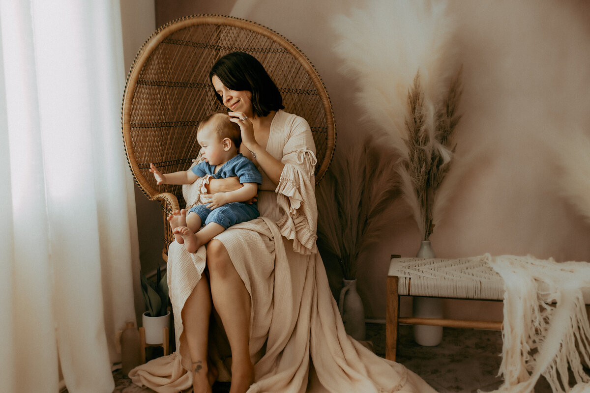 A woman in a beige dress sits on a wicker chair, holding a baby dressed in blue, next to a bench with a white blanket in a softly lit room.