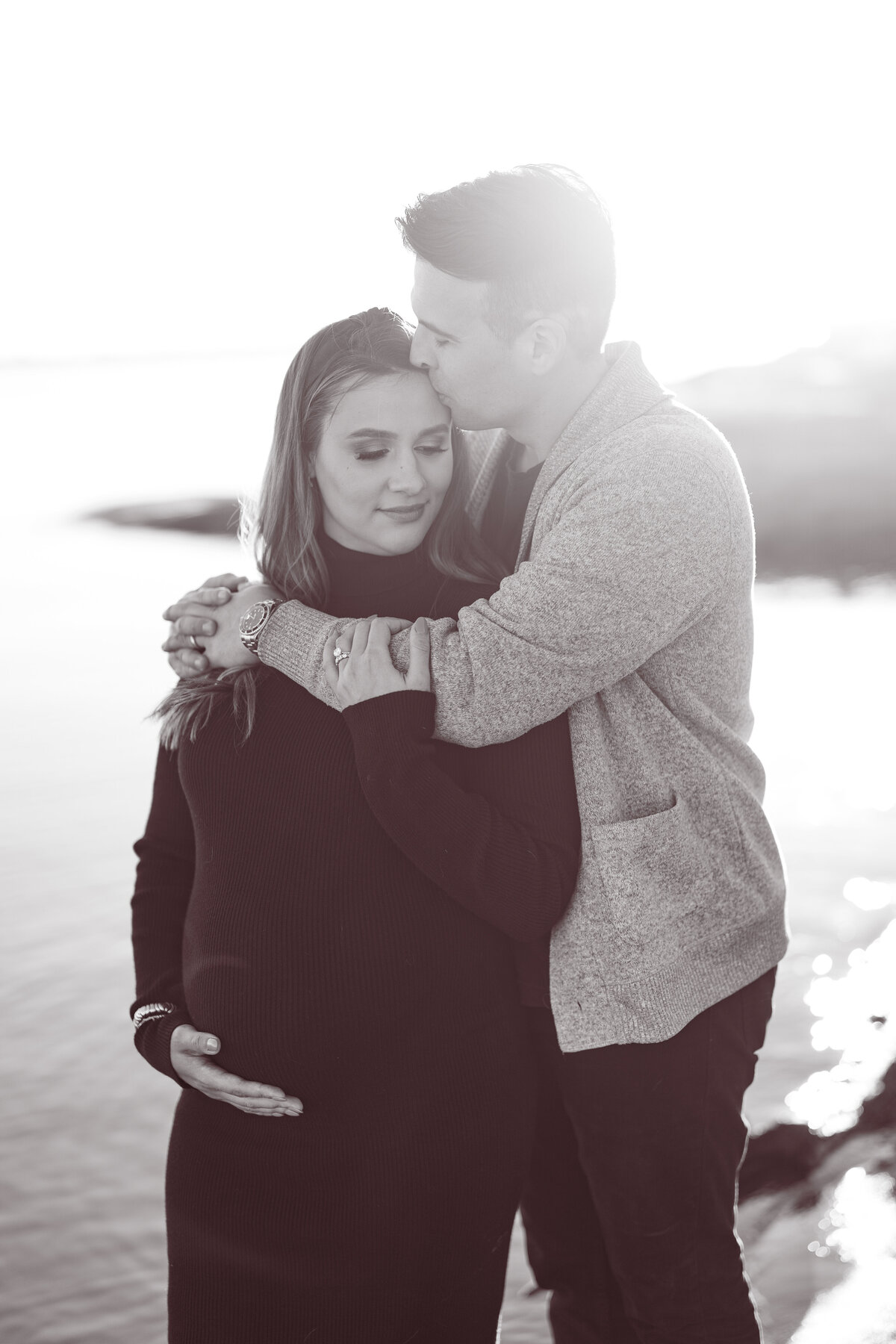 Black and white portrait of a man and a pregnant woman standing holding each other with her hands on her belly next to a lake