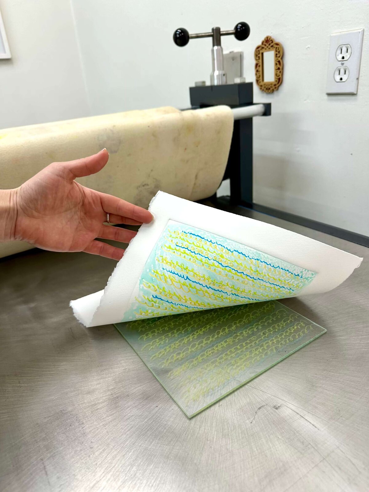 A hand lifts a delicate monoprint of yellow and blue texture marks from a glass plate, revealing soft, painterly results.