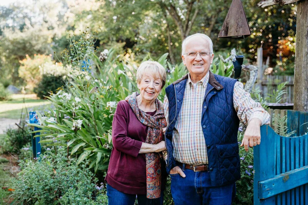 Lovely grandmother and grandfather posing together for a family portrait with a blue fence and greenery surrounding them.