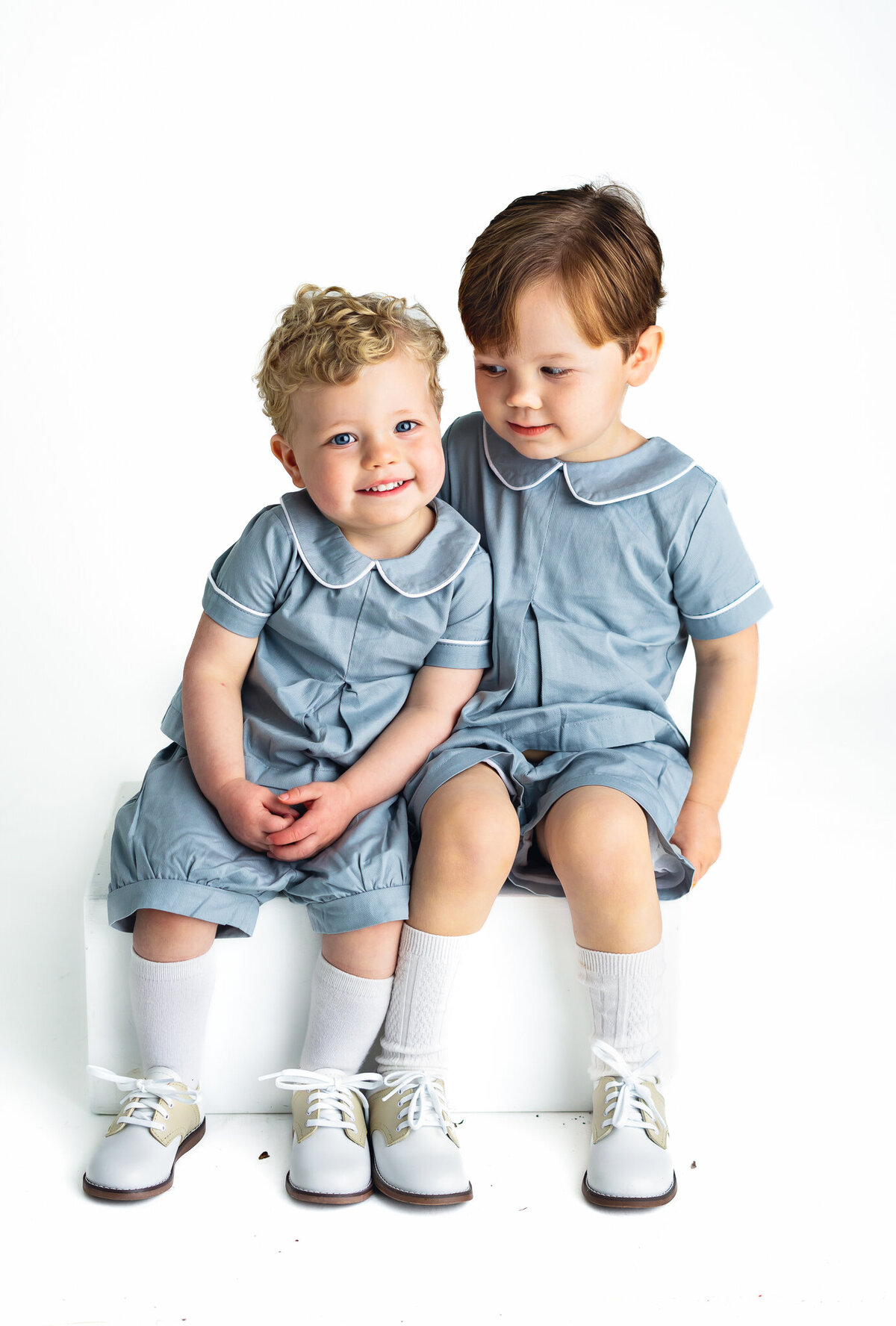 Two young boys in blue outfits are sitting on a white block as they pose for pictures.