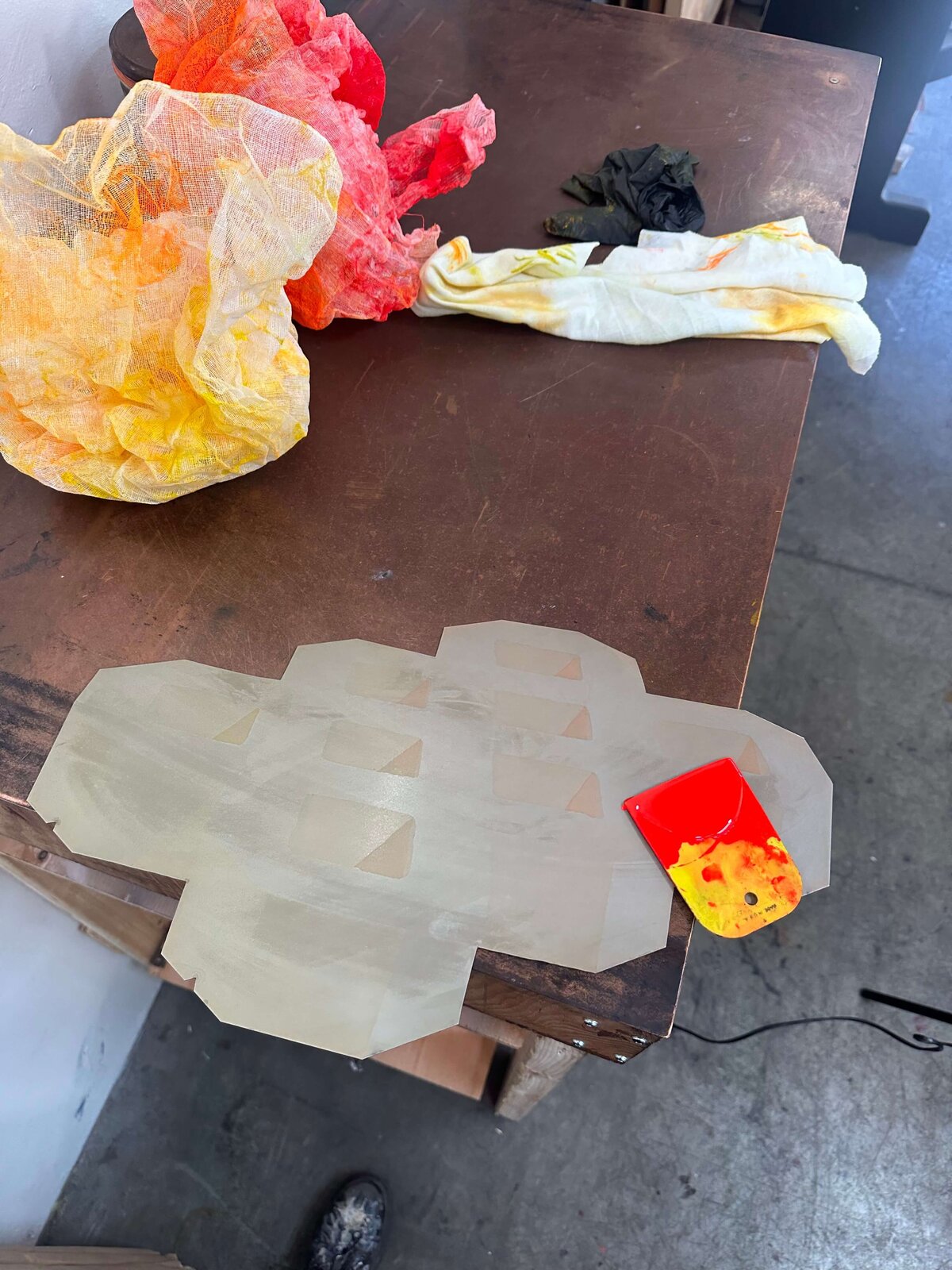 A large copper plate with tonal rectangles prepared for step etching, surrounded by colorful wiping cloths and a red-and-yellow ink spatula.