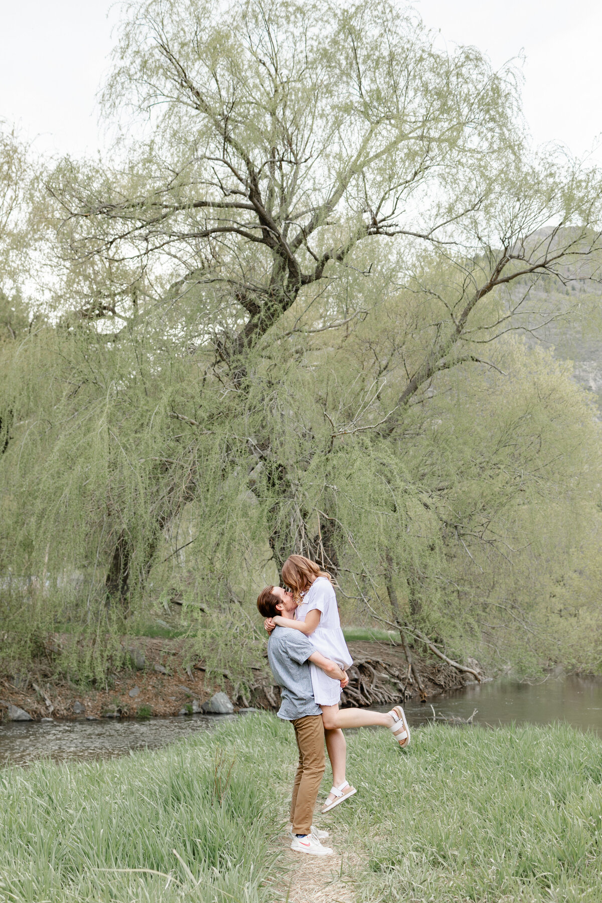 A man gently lifts his fiancée off the ground, holding her close as they laugh together near a peaceful river in Vivian Park, Provo Canyon.