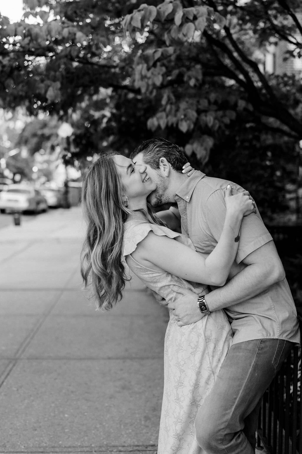 black and white photo of a couple leaning against as fence hugging as one of them kisses the other's neck
