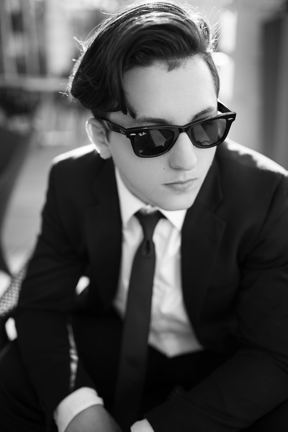 Editorial-style senior portrait of a teenage boy in a black suit and dark horn-rimmed sunglasses, seated outdoors with a confident, iconic look.