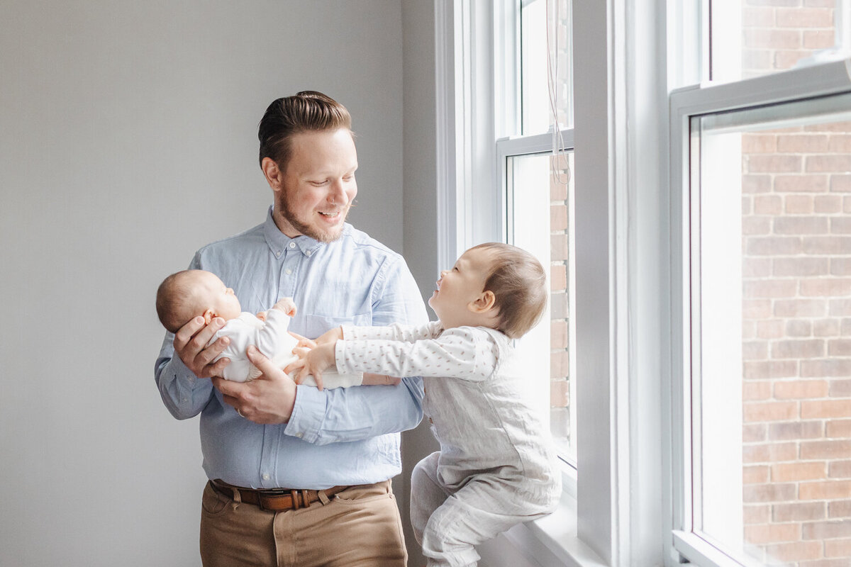 Father holding newborn daughter while smiling at toddler big brother during their at home newborn photography session