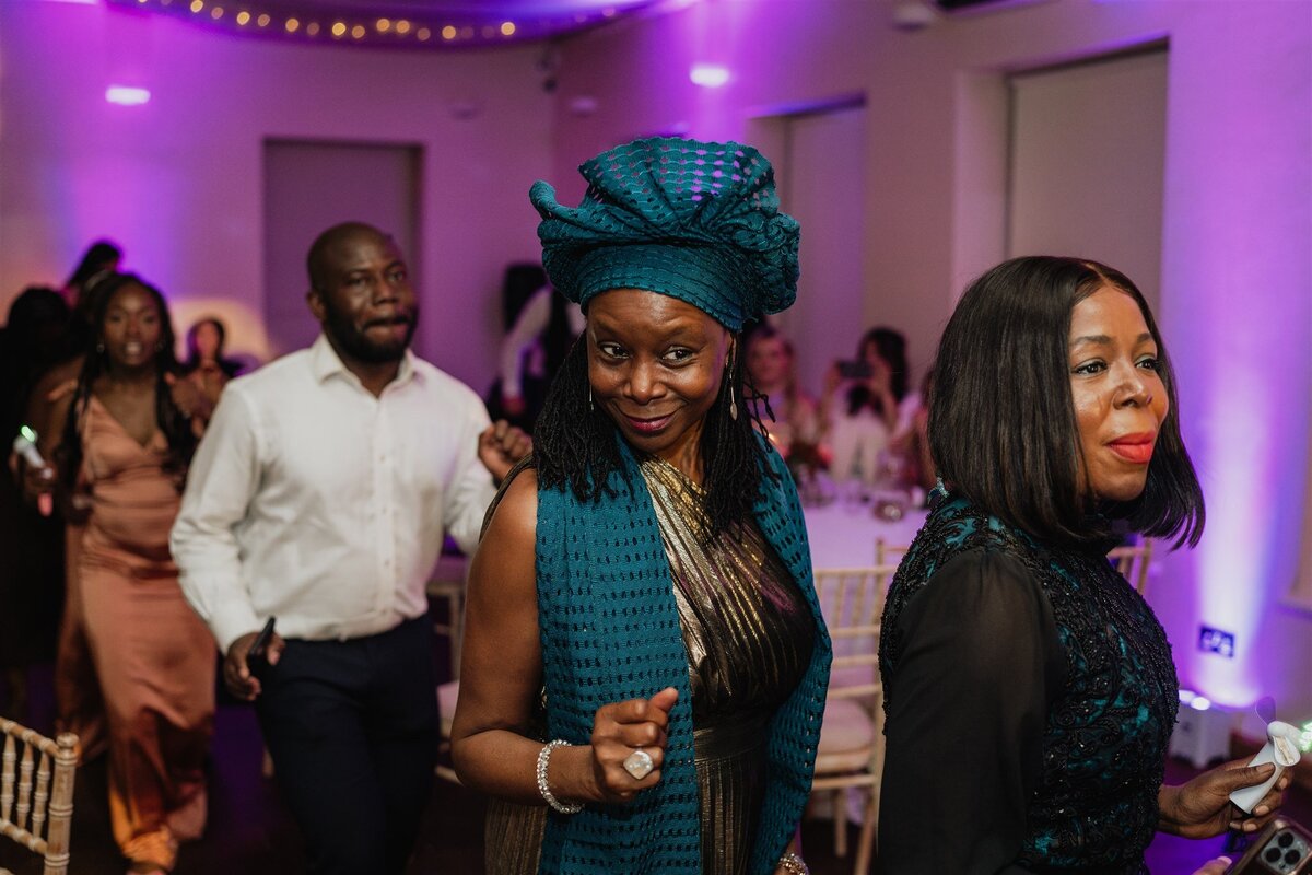 A large group of wedding guests dance in a line and walk through a large venue space. Fairy lights hang around them.