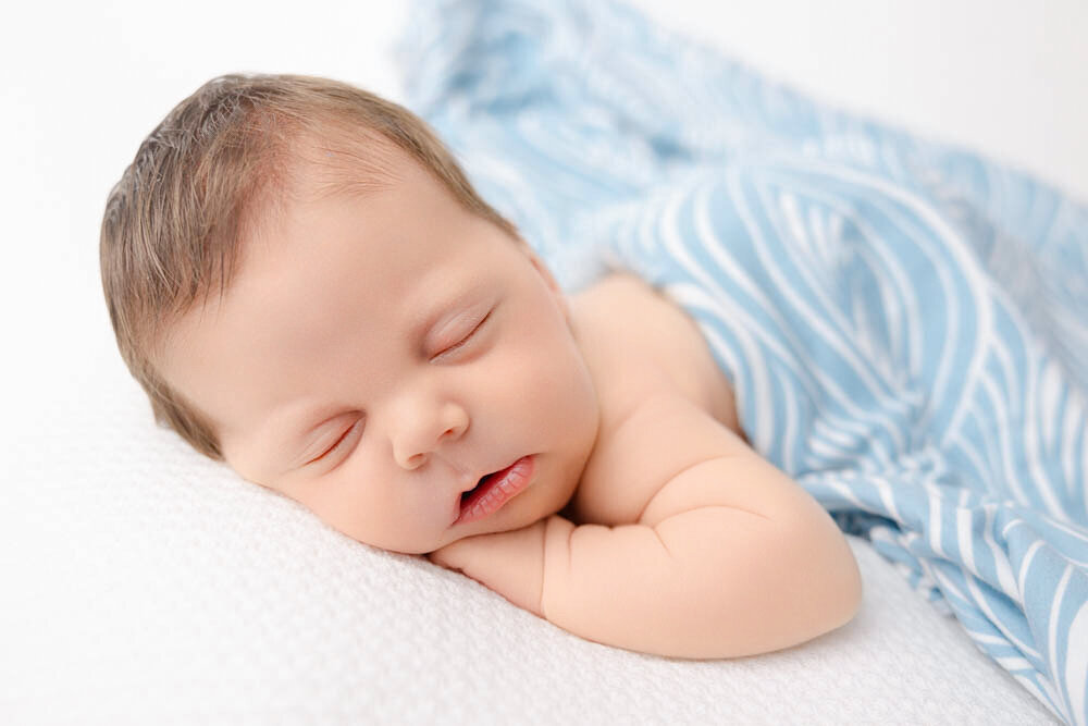 Newborn baby boy lying with hand under chin and covered on his back with a white and blue wave blanket.