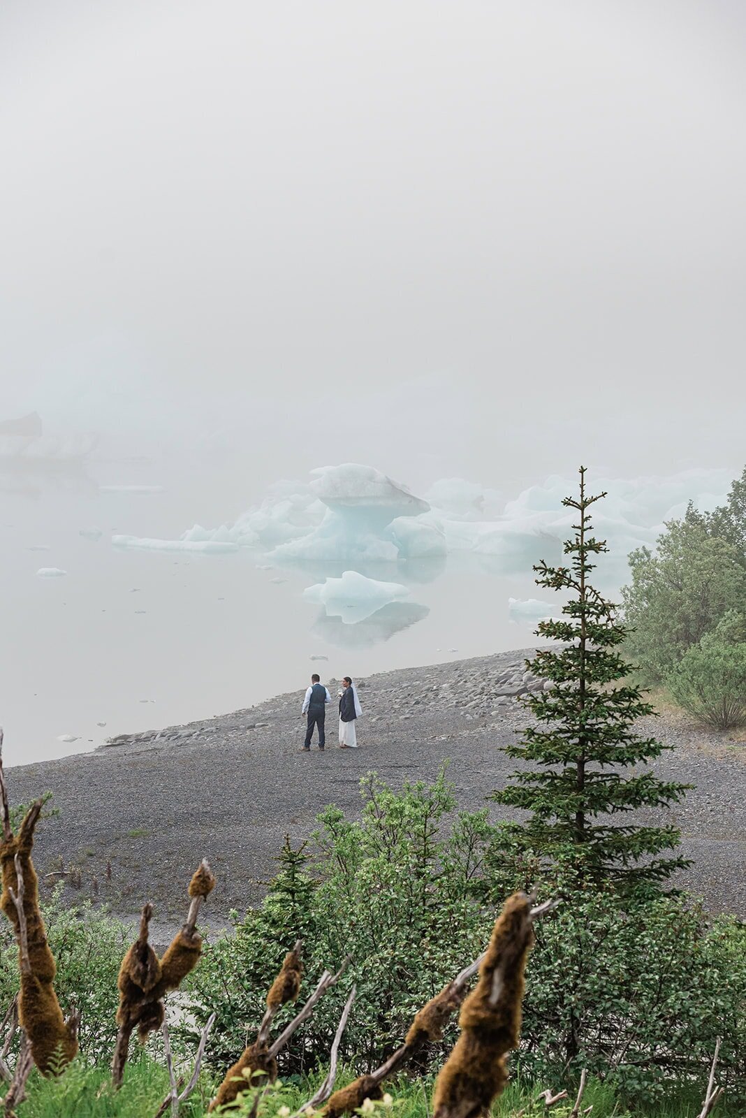 The bride and groom are dressed in their wedding finest, walking across a dark pebbly beach toward the lagoon's edge during their  two day Bear Lake luxury elopement with dogsledding in Alaska. They look small against the large landscape.  Beyond the shore, the background is innumerable blue-tinted white icebergs floating in the lagoon, shrouded in low clouds.   