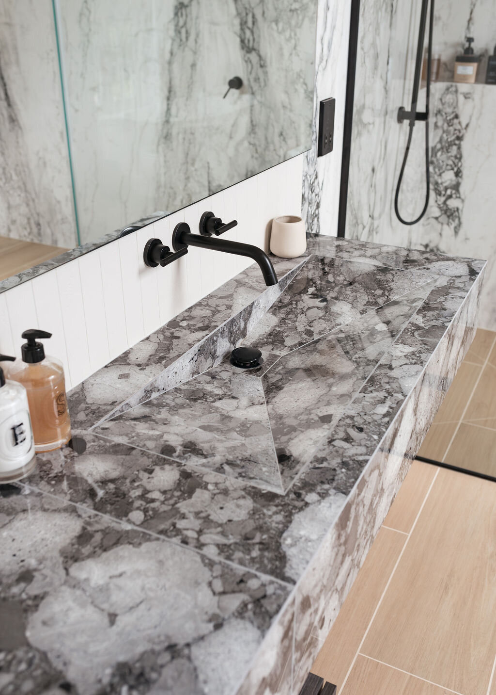 Modern bathroom with a grey marble countertop and black fixtures. A large mirror and glass-enclosed shower with black hardware are visible in the background.