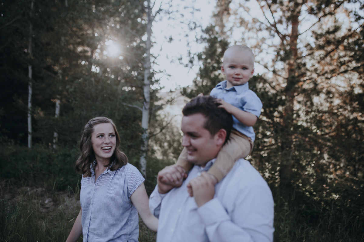 father holding his son on his shoulder as the mother laughs and looks back at the two while they all walk through the woods in the Tetons