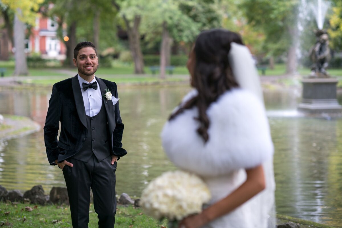 The bride turns her head to look back at the groom, who is smiling from a distance. The image captures a moment of connection and joy, emphasizing the bride's gaze and the groom's engaging expression.