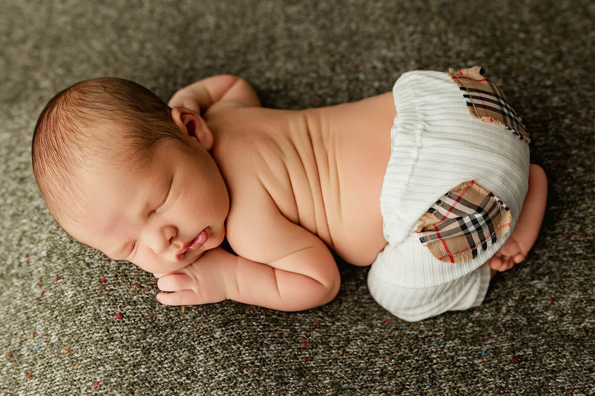 A peaceful newborn baby is curled up sleeping on a textured gray backdrop. The baby has a small tuft of hair and is dressed in a white cloth diaper featuring a patterned back. The scene captures a serene moment, highlighting the innocence and tranquility of early infancy.