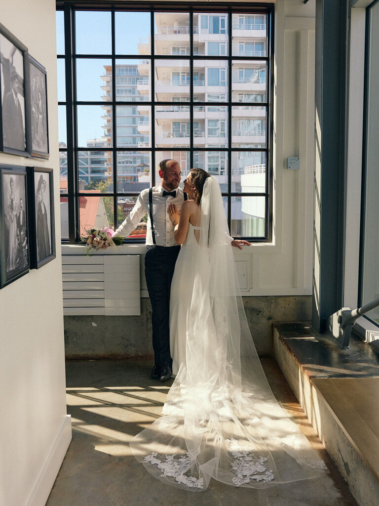 Bride with long cathedral veil hugging groom near the industrial windows of The Wallace in North Vancouver shipyards, featured on the Bronte Bride vendor guide.