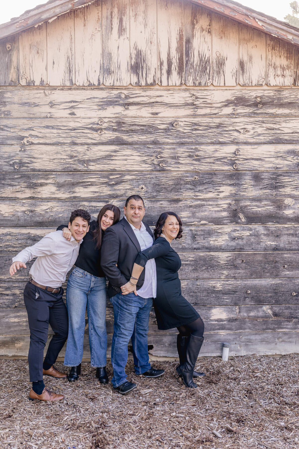 A family of four with dark hair standing in front of a grey wooden barn, laughing and making silly faces at Irvine Ranch Park.